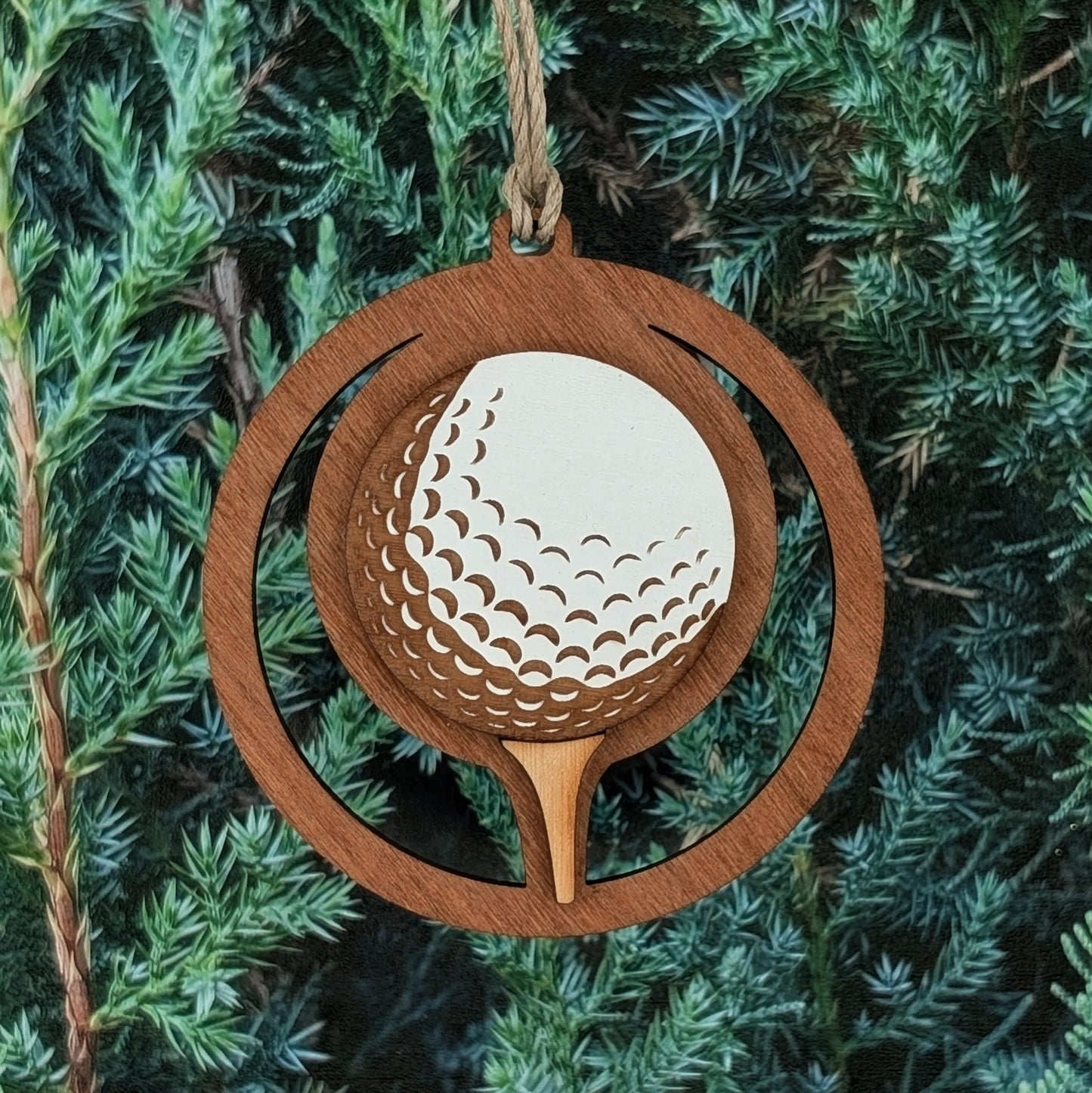 A wooden ornament shaped like a circular frame featuring a golf ball on a tee. The golf ball has carved dimples and is painted white, while the rest of the ornament has a natural stained wood finish. A small hole at the top allows for a rope or string for hanging.