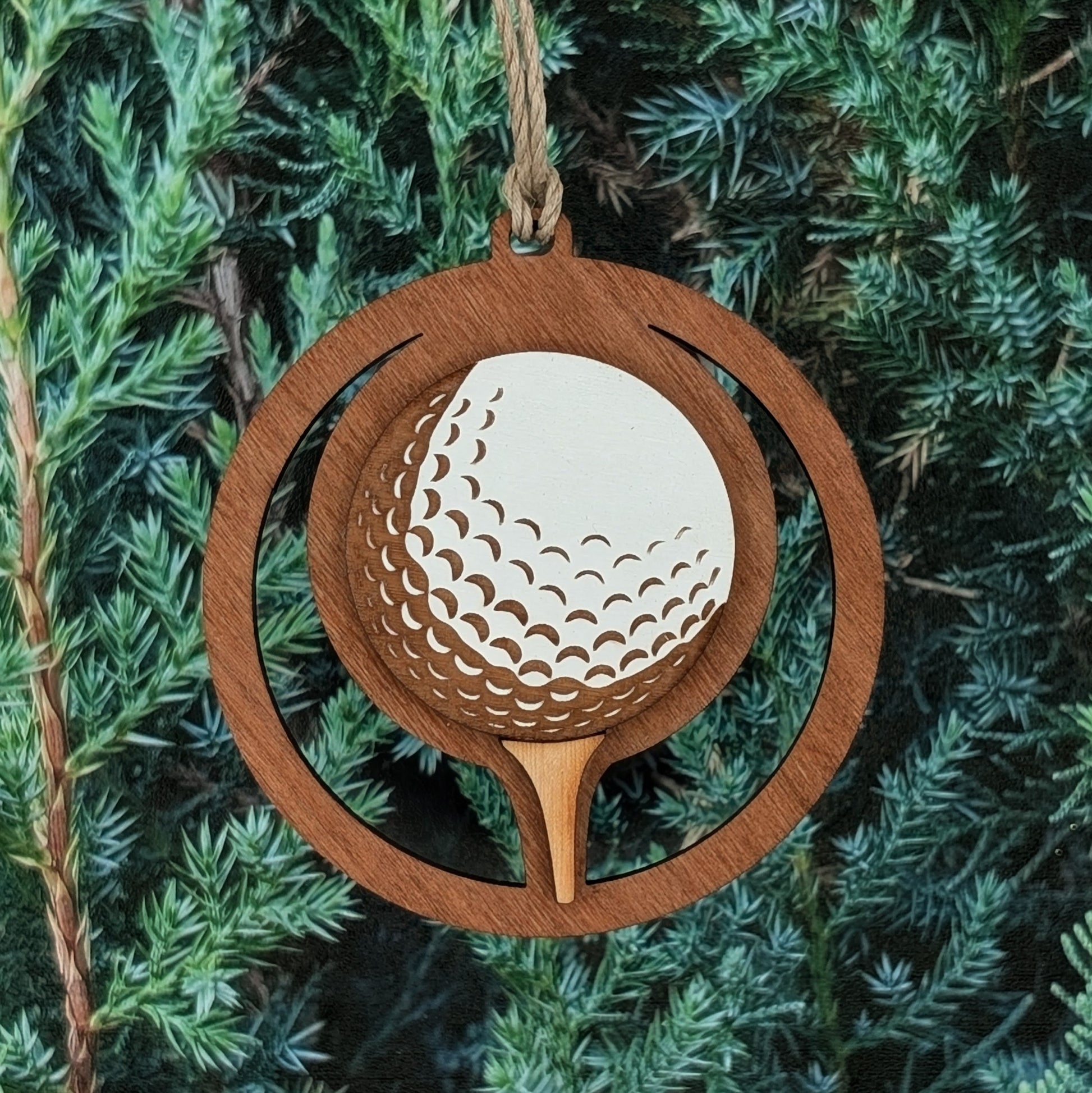 A wooden ornament shaped like a circular frame featuring a golf ball on a tee. The golf ball has carved dimples and is painted white, while the rest of the ornament has a natural stained wood finish. A small hole at the top allows for a rope or string for hanging.